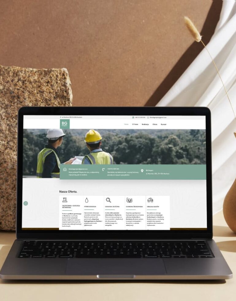 Laptop ustawiony na biurku obok kamienia i wazonów, na ekranie strona BG Project z grafiką dwóch inżynierów w kaskach oraz informacją o ofercie geotechnicznej
